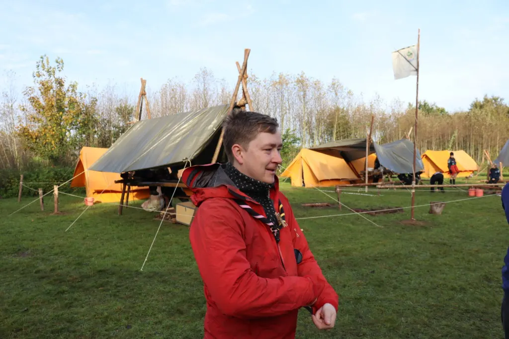 Vejleder i rød jakke på lejrpladsen med telte og flag